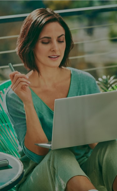 A woman holds an IQOS device while looking at a laptop.